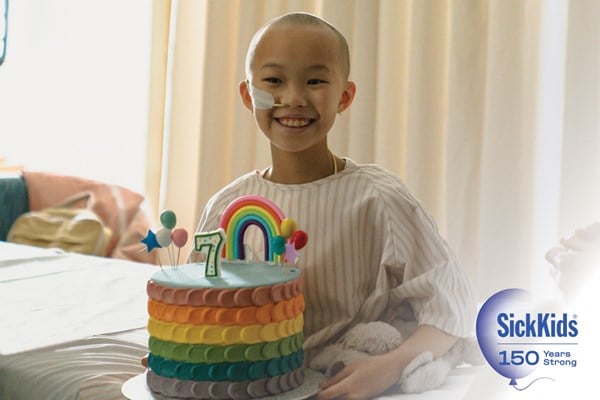 A smiling child holding a rainbow birthday cake with a "7" candle. SickKids 150 Years Strong.