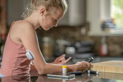 A young girl with diabetes checks her blood sugar levels, a mediation bottle is beside her.