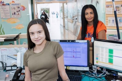 Two people stand on either side of a desk with counter and computers.