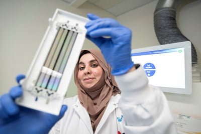 A laboratory professional wearing blue gloves and a white lab coat holds up a piece of laboratory equipment with thin, reflective strips. She appears focused, with a computer screen displaying sequencing-related information in the background.