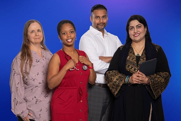 Four people standing together against a blue background. One person wears a light floral top, another wears a red dress with a stethoscope, a third wears a white shirt, and the fourth wears a black and gold patterned outfit holding a tablet.